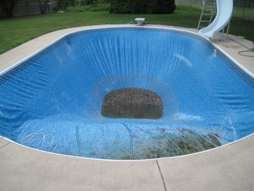 Drained inground pool with a blue vinyl liner, leaves, and standing water in the deep end